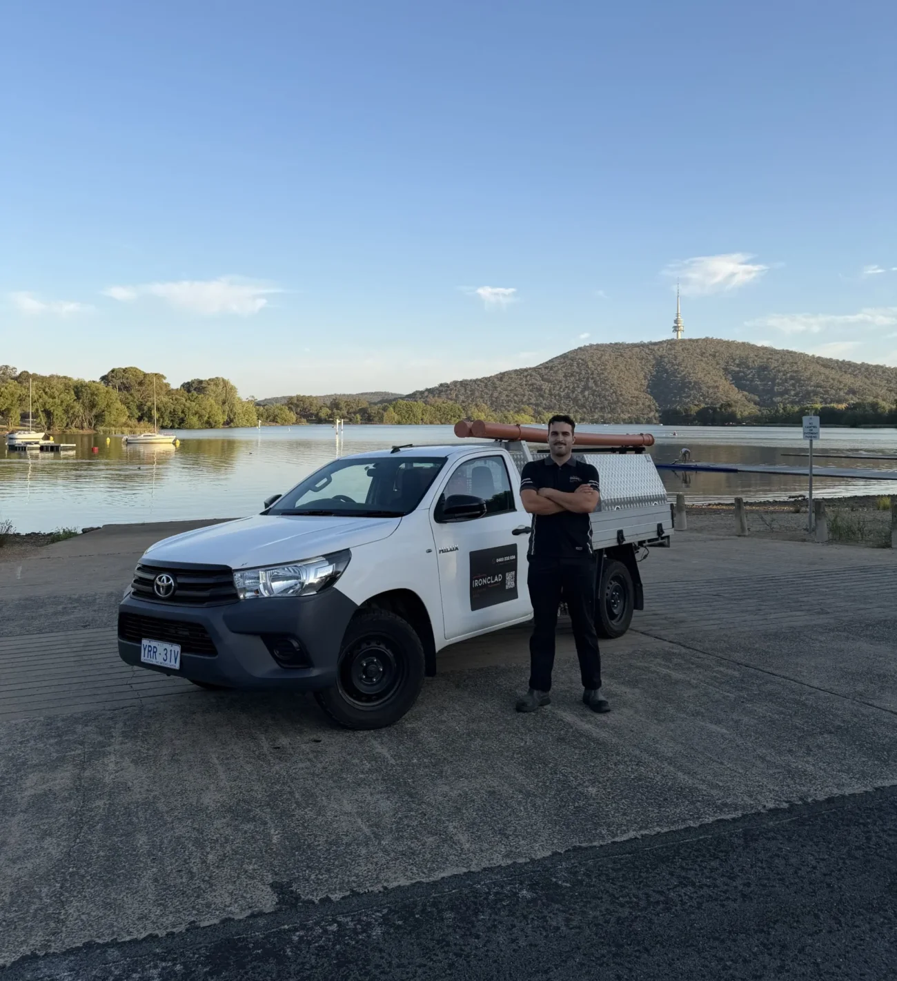 Ben Isaac of Ironclad Plumbing and Gas standing beside his branded work ute at Lake Burley Griffin, with Black Mountain Tower behind